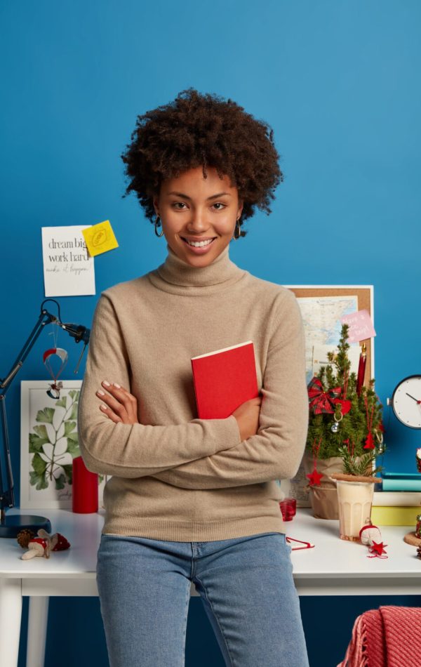 vertical-shot-pleased-curly-haired-female-teacher-prepares-lessons-home-holds-red-textbook-poses-against-desktop